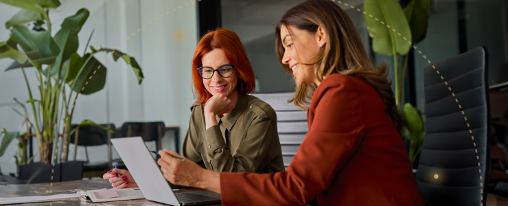 Duas mulheres sentadas à mesa, a trabalhar em conjunto num computador portátil, envolvidas numa discussão e a partilhar ideias.