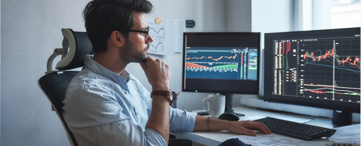 A man sits at a desk with dual monitors, focused on stock market technical analysis and forex trading data.