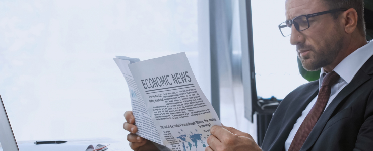 Un homme d'affaires en costume lit un journal intitulé Actualités économiques assis à un bureau avec un ordinateur portable et une plante en arrière-plan.