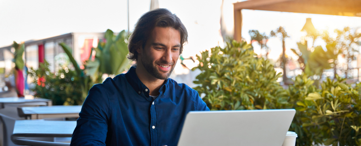 A trader man in a blue shirt smiles while working on a laptop at an outdoor cafe. Greenery surrounds him, and sunlight creates a warm, relaxed atmosphere.