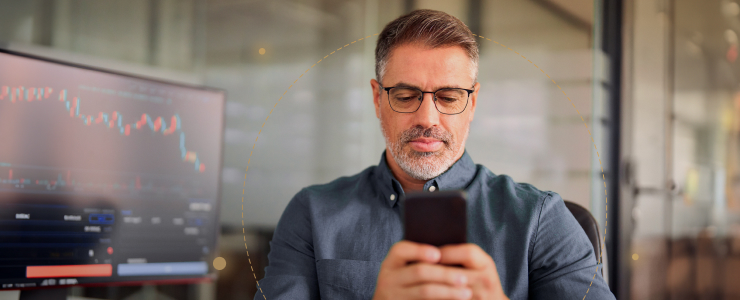 A forex trader man with glasses, focused on a smartphone, sits in an office. A monitor displaying a line graph is in the background. The mood is thoughtful.