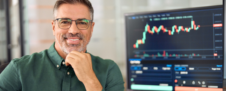 A forex trader, a man in glasses and a green shirt smiles confidently in an office. Behind him, a computer screen displays a vibrant stock trading market chart.