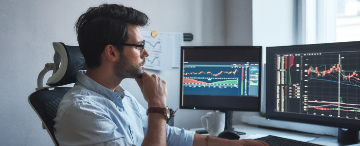 A trader seated at a desk, analyzing stock market graphs on dual monitors in a trading platform setup.