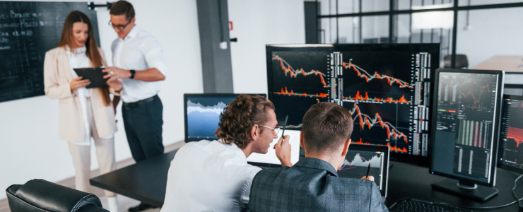 Four people in a modern office; two at desks analyzing financial charts on multiple monitors, and two standing, discussing something on a tablet.