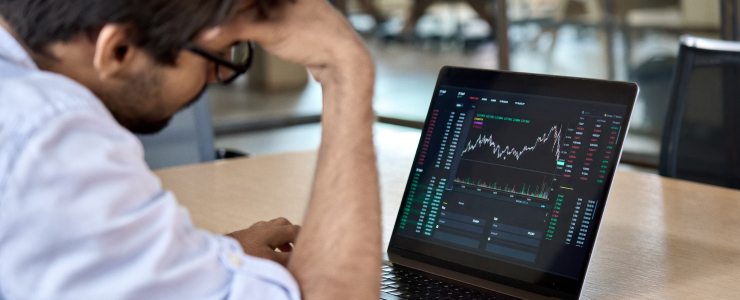 A man sits at a desk, looking stressed while viewing stock market graphs and data on a laptop screen, feeling the emotional rollercoaster of forex trading.