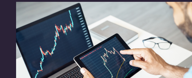 A person using a tablet to analyze stock market charts, with similar graphs displayed on a laptop. The setting is an office, suggesting a business context.
