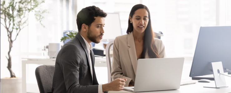 Two business professionals looking at a laptop and collaborating in a modern office, learning how to choose a beginner forex broker.