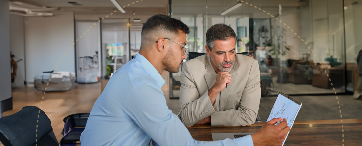 Two men discussing financial data in a bright office, with one pointing at a graph on a tablet while reviewing risk management in forex strategies.